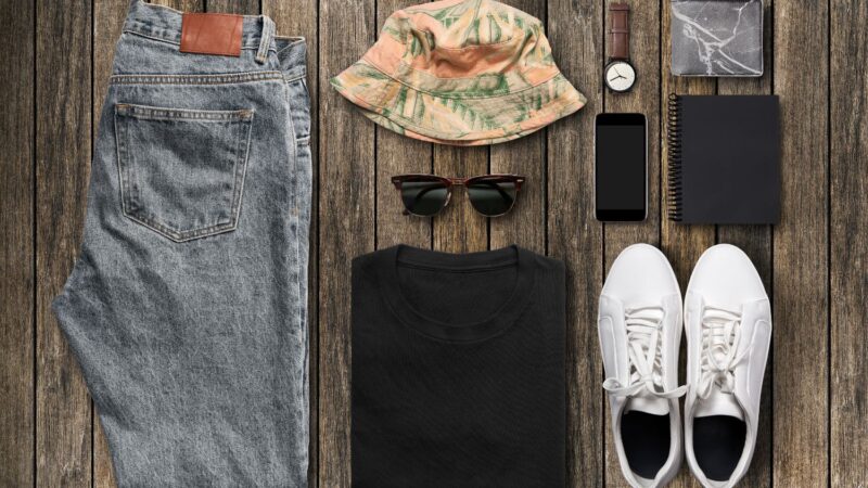 Image of folded clothing, including a pair of blue jeans, a summer hat, sunglasses, a black t-shirt, white trainers, and accessories.